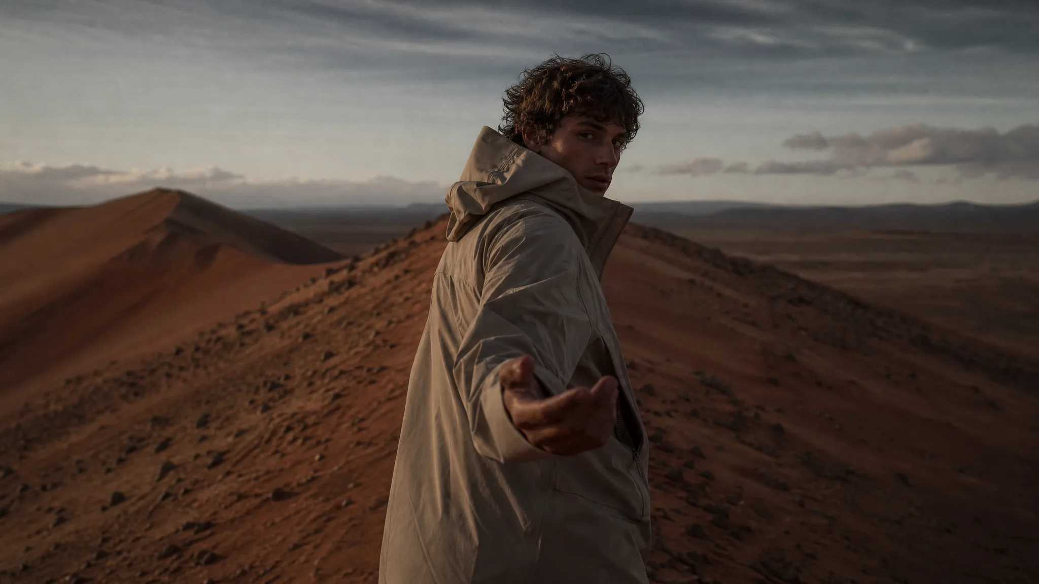 Man on a desert dune ridge at dusk looking back and beckoning the viewer to follow