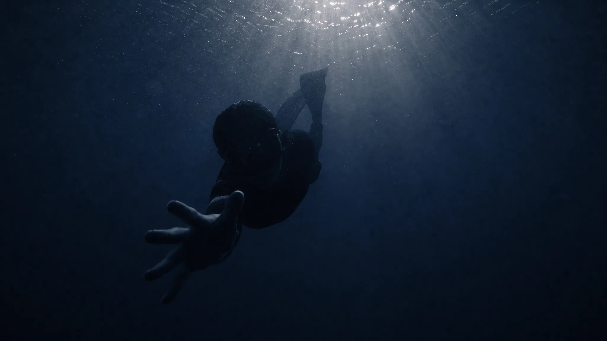 Freediver silhouette in deep blue water reaching an open hand toward the camera