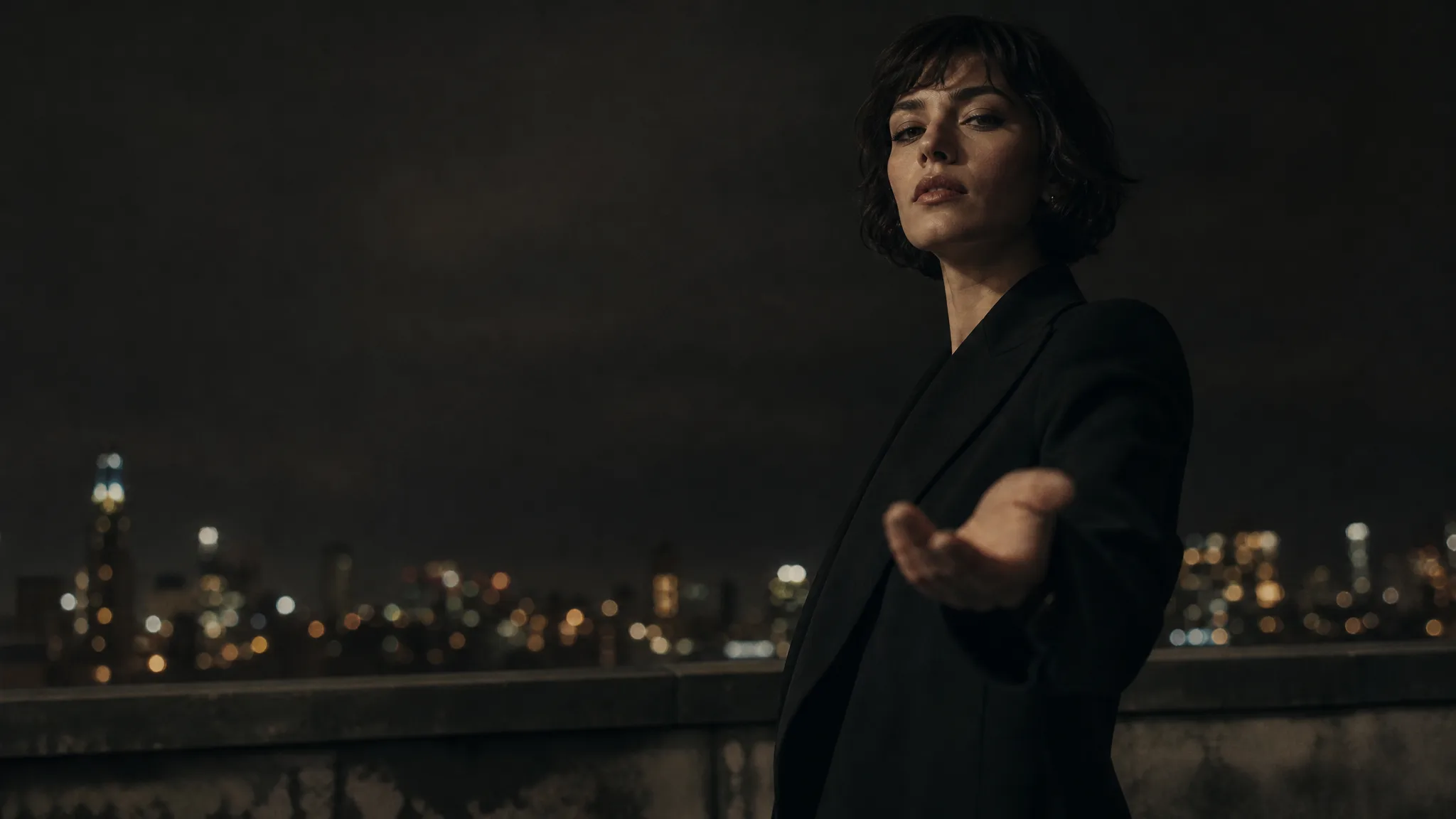 Woman in a tailored black coat on a night rooftop holding an open hand out toward the camera