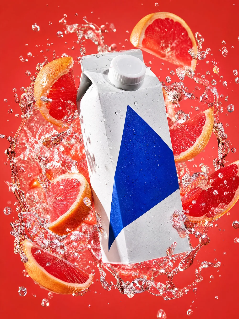 Karton minuman putih dengan panel biru melayang di tengah lingkaran irisan grapefruit dan percikan cairan.